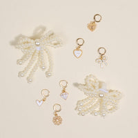 Two pearl bow hair clips and seven gold and white heart, bow, flower, and diamond-shaped charms from the-darling-effect’s Pretty in Pearl Dazzle Shoe Charm Pack are arranged on a light beige surface.