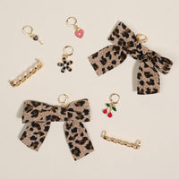 The Darling Effect's Wild & Free Dazzle Shoe Charm Pack includes two leopard print bows, two gold hair clips, five small gold charms (heart, flower, cherries, pearl), and sneaker charms—arranged on a light background.