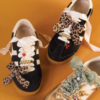 Three sneakers on an orange surface, styled with The Darling Effect’s Wild & Free Dazzle Shoe Charm Pack: black pair with leopard bows, gold chains, cherry charms; gold pair with green gingham bows and assorted gold charms.