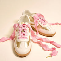 A pair of white and gold sneakers with light brown soles is displayed on a light background, featuring The Darling Effect's Frilly Pink Step It Up Shoelaces.