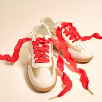 A pair of white and gold sneakers with gum soles, featuring bright red, wide, durable shoelaces loosely laced and spread out on a light background.