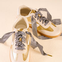 A pair of white and gold sneakers with The Darling Effect's Gingham Black Step It Up Shoelaces, shown on a cream-colored surface.