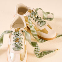 A pair of cream and gold sneakers with The Darling Effect's Gingham Green Step It Up Shoelaces, featuring a green and white gingham pattern, displayed on a light beige surface.