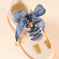 A gold and cream sneaker with The Darling Effect's Gingham Navy Step It Up Shoelaces, featuring a blue and white gingham pattern tied in a bow.