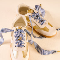 A pair of cream and gold sneakers laced with The Darling Effect's Gingham Navy Step It Up Shoelaces is displayed on a light beige surface.