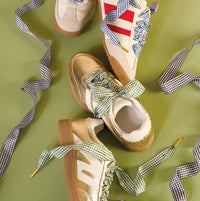 Three pairs of stylish sneakers with gum soles and patterned laces—two with The Darling Effect's Gingham Pink Step It Up Shoelaces, one with red stripes and blue-and-white laces—are displayed on a green background.
