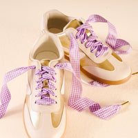 Two cream and gold sneakers with gum soles are shown on a light surface, featuring Gingham Purple Step It Up Shoelaces by The Darling Effect—wide, purple and white checkered laces loosely tied for a stylish touch.