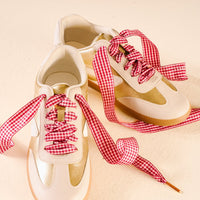 A pair of white sneakers with gold accents, laced with The Darling Effect's Gingham Red Step It Up Shoelaces, displayed on a light cream-colored surface.