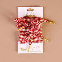 Add a pop of style to your shoes with the Gingham Red Step It Up Shoelaces from The Darling Effect—red and white gingham laces with gold aglets, tied in bows and displayed on a branded card against a beige background.