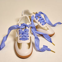 A pair of white and gold sneakers with brown soles, laced with The Darling Effect's Gingham Royal Blue Step It Up Shoelaces, are placed side by side on a plain, light-colored surface.