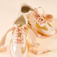 A pair of white and gold sneakers with The Darling Effect's Gingham Tan Step It Up Shoelaces, featuring a pink and white gingham pattern, are placed side by side on a light beige surface with the durable laces loosely arranged around them.