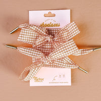 Two beige and white gingham shoelaces, tied in bows, are displayed on a pink card labeled "Gingham Tan Step It Up Shoelaces" by The Darling Effect, featuring the message "Accessorize Your Shoes.