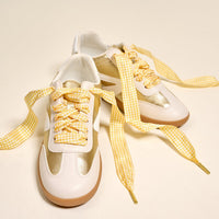 A pair of white sneakers with metallic gold accents and The Darling Effect's Gingham Yellow Step It Up Shoelaces are placed on a light beige surface.