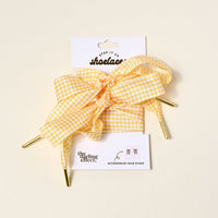 Gingham Yellow Step It Up Shoelaces by The Darling Effect feature yellow and white gingham with gold aglets, tied in a bow on packaging labeled with the brand name and product, plus a sneaker illustration at the bottom.