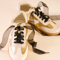 White and gold sneakers with Sheer Black Step It Up Shoelaces by The Darling Effect and gum soles, shown as a pair on a cream surface, for a chic modern style.