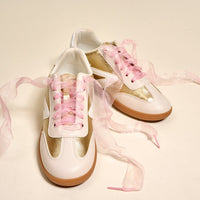 A pair of white and gold sneakers featuring The Darling Effect's Sheer Pink Step It Up Shoelaces are displayed on a light background, with the translucent pink laces from the brand loosely arranged around the shoes.