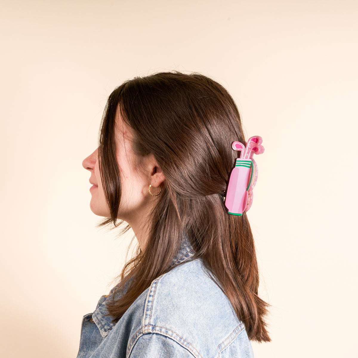 A woman with brown hair in profile wears a denim jacket and styles her hair with The Darling Effect’s Golf Clubs Shape Up Claw Clip—a pink, golf bag–shaped accessory—against a plain, light background.
