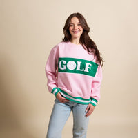A woman with long brown hair wears The Darling Effect Golf Signature Crew sweatshirt in pink and green with light blue jeans, smiling against a plain beige background.