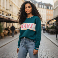 A woman with long curly hair wears The Darling Effect's Mahj Signature Crew, featuring “MAHJ” on the front, while standing on a cobblestone street in a European city with charming buildings and outdoor cafes.