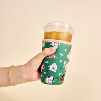A hand holds an iced coffee in a clear cup wrapped with The Darling Effect’s Large-Dog Days Sip Savvy Drink Sleeve, featuring cute dog, bone, and tennis ball designs. The person is also wearing a gold beaded bracelet.