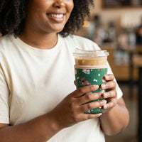 A woman smiles, holding an iced coffee in The Darling Effect's green cup with colorful animal prints and the Large-Dog Days Sip Savvy Drink Sleeve. She wears a light t-shirt and stands in a cafe.