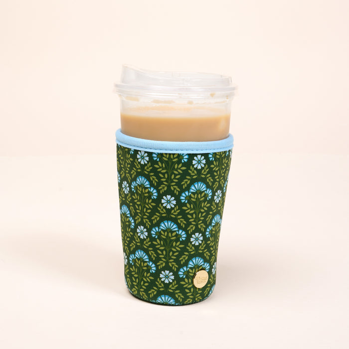 The Darling Effect's Large-Everbloom Forest Green Sip Savvy Drink Sleeve, featuring a blue floral pattern and light blue trim, wraps around an iced coffee to keep it cool and stylish.