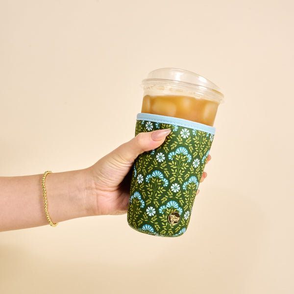 A hand with a gold beaded bracelet holds an iced coffee in The Darling Effect's Large-Everbloom Forest Green Sip Savvy Drink Sleeve against a cream background.