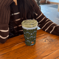 A disposable cup with a green beverage features The Darling Effect Large-Everbloom Forest Green Sip Savvy Drink Sleeve and rests on a wooden table. A person wearing a striped sweater is partially visible.