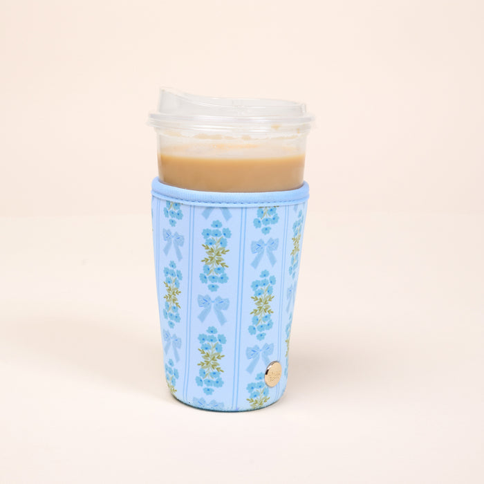 A plastic cup of iced coffee with a clear lid is wrapped in The Darling Effect's Large-Oh So Fancy Blue Sip Savvy Drink Sleeve, featuring blue flowers and ribbon patterns, set against a plain, light background.