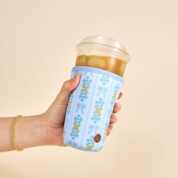 A hand holds an iced drink in a clear cup with a domed lid, wrapped in The Darling Effect's Large-Oh So Fancy Blue Sip Savvy Drink Sleeve. The background is plain beige.