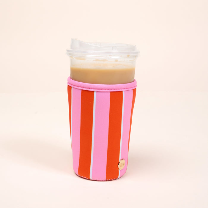 A clear plastic cup with iced coffee and a lid is wrapped in The Darling Effect's Large-Sicily Stripes Pink Sip Savvy Drink Sleeve, featuring red and pink stripes with a gold label, set against a light beige background.