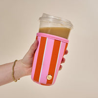 A hand wears a gold beaded bracelet and holds a clear cup with a tan drink, inside The Darling Effect Large-Sicily Stripes Pink Sip Savvy Drink Sleeve featuring pink and orange stripes and a gold button.