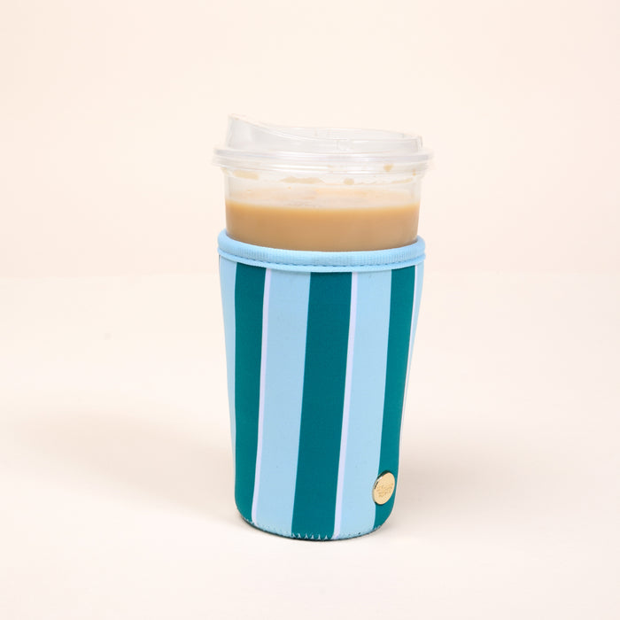 A plastic cup of iced coffee with a clear lid is wrapped in The Darling Effect's Large-Sicily Stripes Teal Sip Savvy Drink Sleeve, featuring blue and teal stripes, and sits on a plain light-colored surface with a neutral background.
