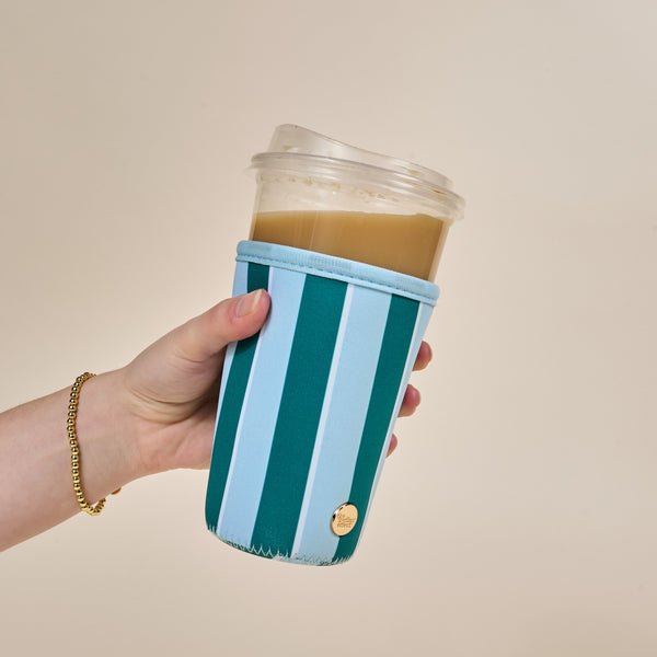 A hand holds an iced coffee in a clear cup with The Darling Effect’s Large-Sicily Stripes Teal Sip Savvy Drink Sleeve; the hand wears a gold bracelet and the background is plain light beige.