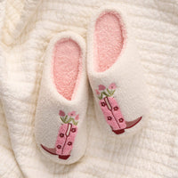 The Darling Effect’s Cowgirl Boots Fuzzy Slippers feature a plush white exterior, pink interior, and embroidered pink cowboy boots with flowers—perfect for lounging on a cream quilted blanket.