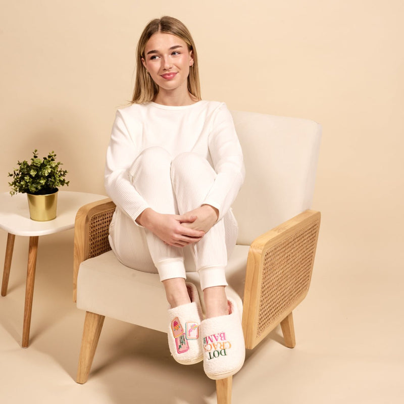 A woman in white loungewear sits on a beige chair, smiling with her arms on her knees. She wears The Darling Effect Dot Crack Bam Fuzzy Slippers—one features a drink illustration, the other reads “Dog Hair Don’t Care.” A plant is nearby.