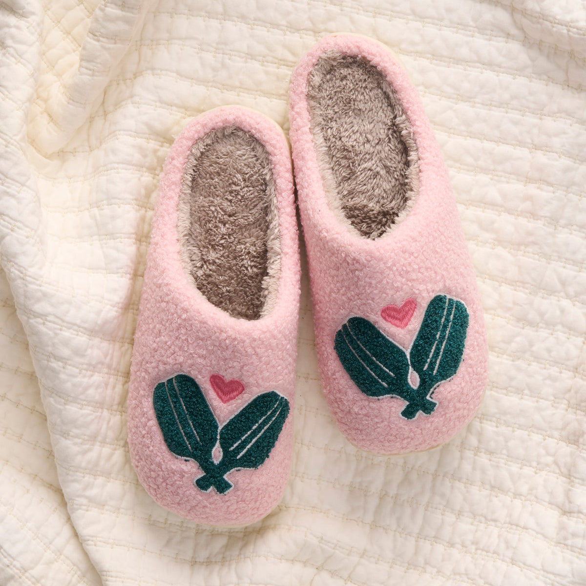 The Darling Effect's Pickleball Paddles Fuzzy Slippers, pink with gray interiors and green cactus and pink heart designs, are displayed on a white quilted blanket.
