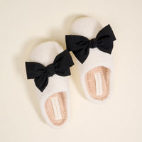 The Darling Effect's Betty Bow Black Fuzzy Slippers feature plush white material and black bows, displayed against a cream background.