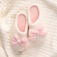 The Darling Effect’s Betty Bow Blush Fuzzy Slippers, featuring white fuzz, pink insoles, and large pink bows, rest on a textured cream blanket for a cozy, charming touch.