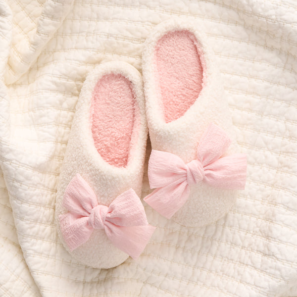 The Darling Effect’s Betty Bow Blush Fuzzy Slippers, in fluffy white with large pink bows, rest on a soft quilted white blanket.