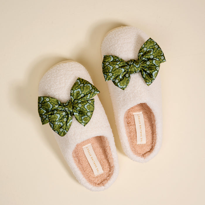 The Darling Effect’s Betty Bow Everbloom Forest Green Fuzzy Slippers feature a cozy cream exterior, soft pink inner lining, and green bows with a white floral pattern, shown on a light beige background.
