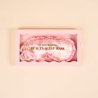 A wavy-edged pink box holds The Darling Effect’s Oh So Fancy Pink Beauty Sleep Mask—a light pink satin mask with a floral pattern and gold text: THE DARLING EFFECT and BEAUTY SLEEP MASK.
