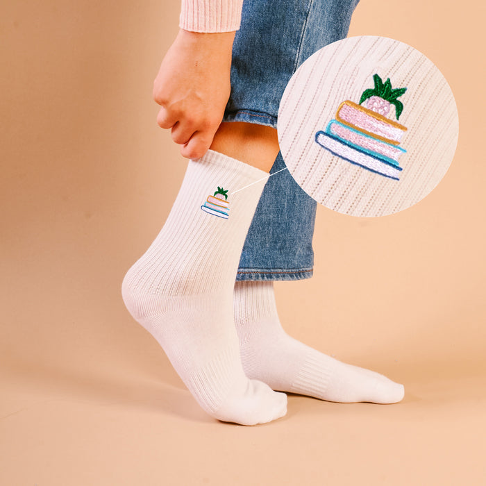 A person wears Book Nook Embroidered Crew Socks in white, featuring a layered cake with a strawberry on top. A close-up inset shows the vibrant cake embroidery. The background is soft beige.