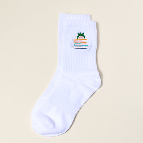 The Book Nook Embroidered Crew Sock is a single white women’s sock with a pineapple perched on three books design—ideal for those who love unique, colorful socks.