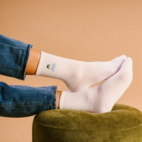 Someone wearing Book Nook Embroidered Crew Socks with a small design and blue jeans relaxes, feet up on a green ottoman, set against a beige background.