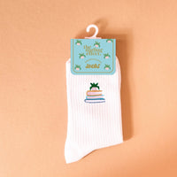 The Book Nook Embroidered Crew Socks is a white ribbed women’s sock featuring embroidered books and a small green plant, shown against a peach background with packaging labeled “the darling effect” on a light blue tag.