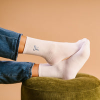 A person wearing The Darling Effect's Blue Bow Embroidered Crew Socks rests their feet on a green ottoman, dressed in blue jeans against a light tan background.