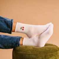 A person in blue jeans rests their feet on an olive green stool, wearing The Darling Effect’s Cherries Embroidered Crew Socks against a soft beige background—perfect for fans of stylish women’s socks.