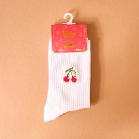 The Darling Effect's Cherries Embroidered Crew Socks, featuring a white ribbed design with cherry embroidery, are shown on a light brown background. The women's socks come packaged with a pink label decorated with yellow text and cherry illustrations.