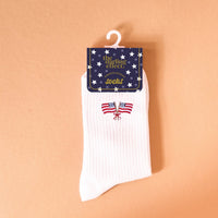 Add a patriotic flair to any outfit with the American Flags Embroidered Crew Socks by The Darling Effect—featuring white fabric adorned with two crossed American flags, and packaged in a star-patterned label set against a peach background.